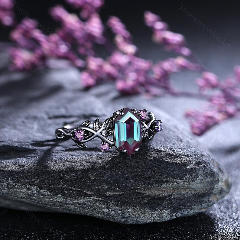 May include: A silver ring with a large, emerald-cut, colour-changing gemstone. The ring features a floral design with small pink gemstones and leaf accents. The ring is set on a dark grey stone with blurred purple flowers in the background.