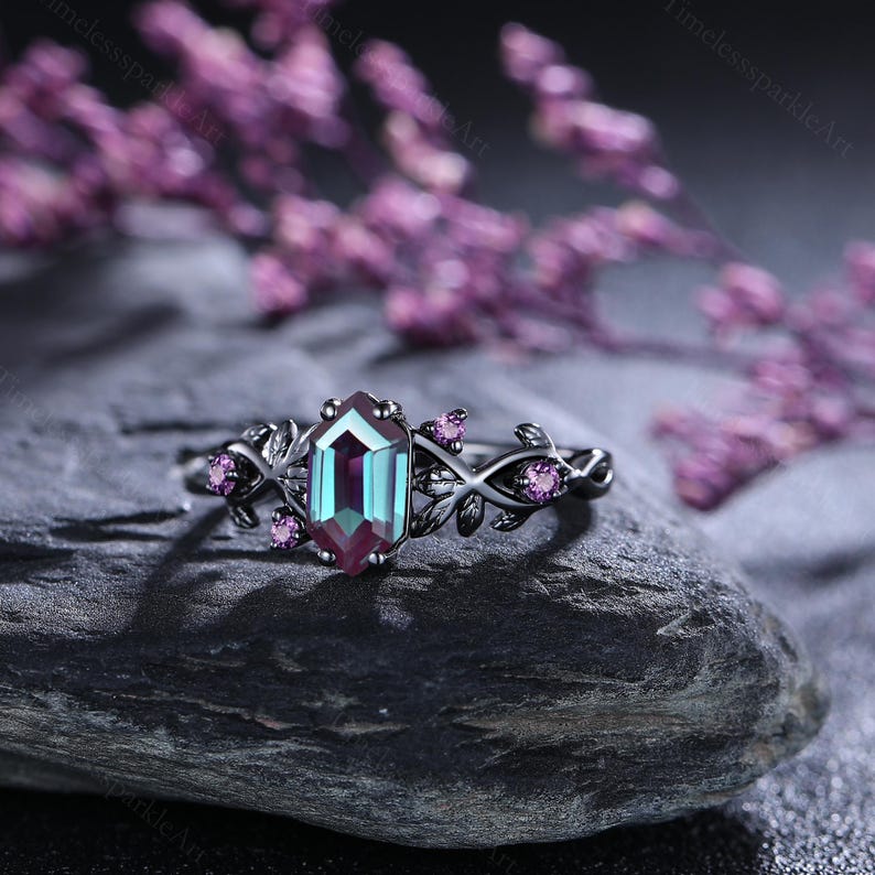 May include: A silver ring featuring a large, rectangular gemstone that changes colour. The gemstone shifts from teal to purple. The ring has a floral design with small pink gemstones and leaves. The ring is set on a dark grey stone.