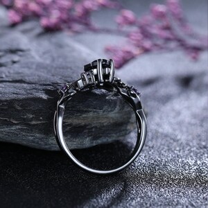 May include: A black ring featuring a dark square central stone and small purple accent stones. The ring has an intricate, ornate design and is set against a dark, textured backdrop. The ring's band curves elegantly, highlighting the detailed metalwork.