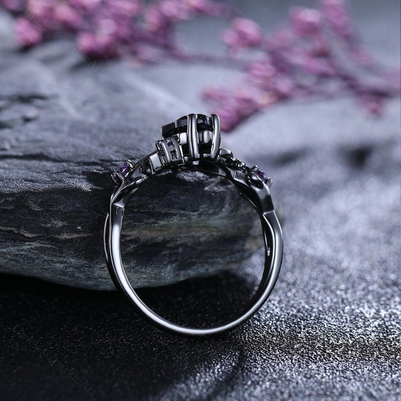 May include: A black ring featuring a dark square central stone and small purple accent stones. The ring has an intricate, ornate design and is set against a dark, textured backdrop. The ring's band curves elegantly, highlighting the detailed metalwork.