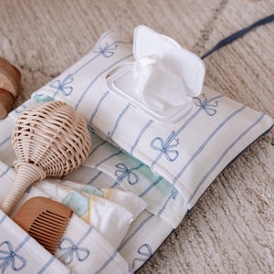 May include: A white and blue striped diaper bag with a blue ribbon pattern. The bag has a pocket for a pack of wipes, a pocket for a rattle, and a pocket for diapers.