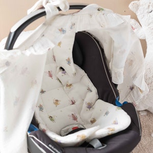 May include: A black car seat with a white fabric cover featuring a fairy pattern. The cover has a hood that can be pulled over the seat.