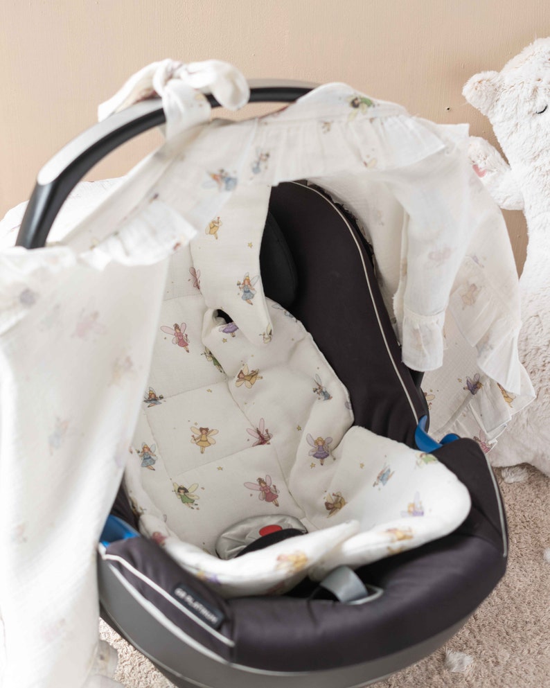 May include: A black car seat with a white fabric cover featuring a fairy pattern. The cover has a hood that can be pulled over the seat.