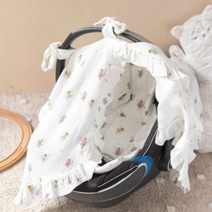 May include: A white muslin baby car seat cover with a fairy print. The cover has a ruffle trim and ties at the top.