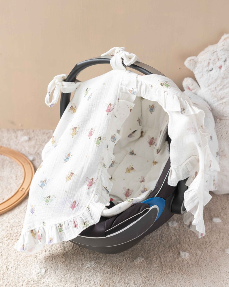 May include: A white muslin baby car seat cover with a fairy print. The cover has a ruffle trim and ties at the top.
