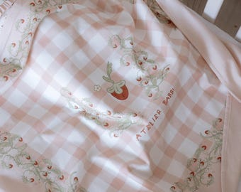 Può includere: Una copertina per neonati in gingham rosa e bianco con un motivo a fragola e fiori. La copertina ha un bordo bianco con il testo "Atelier Babbi" in verde.