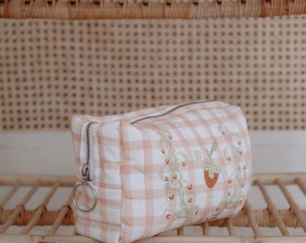 Può includere: Una trousse da trucco a quadri rosa e bianchi con un ricamo floreale che presenta fragole. La trousse ha una cerniera argentata e un anello di trazione argentato.