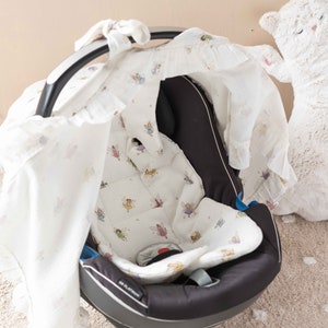 May include: A black baby car seat with a white and light pink fairy-themed cover. The cover has a ruffle trim and a hood that can be pulled over the seat. The car seat is sitting on a light brown carpet.