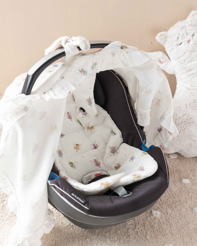 May include: A black baby car seat with a white and light pink fairy-themed cover. The cover has a ruffle trim and a hood that can be pulled over the seat. The car seat is sitting on a light brown carpet.