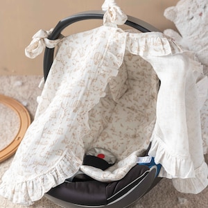 May include: A white and beige baby car seat cover with a ruffled edge and a floral pattern. The cover is made of a soft, breathable fabric and is designed to provide comfort and protection for babies.