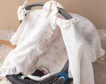 May include: A white baby car seat cover with pink bows printed on it. The cover is made of a soft, breathable fabric and has a ruffled edge. The cover is designed to protect the car seat from dirt and spills.