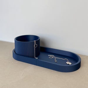 Può includere: Un vassoio portagioie blu navy con un supporto cilindrico e un vassoio ovale. Il vassoio ha una texture a coste e contiene due collane d'argento. L'organizer per gioielli è un modo elegante per riporre collane, anelli e altri accessori.