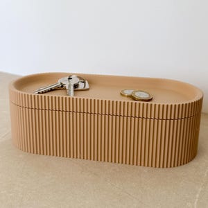 Könnte beinhalten: Eine beige, ovale Aufbewahrungsbox mit geriffelter Außenseite. Die Box enthält einen Satz Schlüssel und zwei Münzen. Das Design ist minimalistisch und modern.