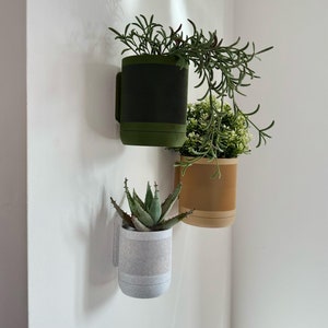 Pode incluir: Três vasos de parede com vegetação. O vaso superior é verde-oliva com uma faixa texturizada verde-escura e uma alça. O vaso do meio é bege com um design de chevron. O vaso inferior é cinza claro com uma faixa texturizada.