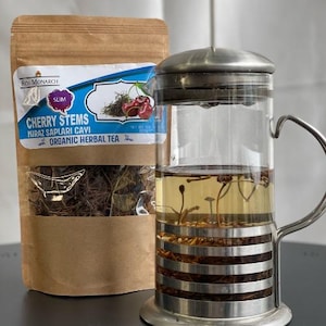 May include: A brown paper bag with the text "Cherry Stems" and "Organic Herbal Tea" on it. The bag contains dried cherry stems. A glass tea infuser with a metal lid and handle is filled with a light brown liquid and cherry stems.