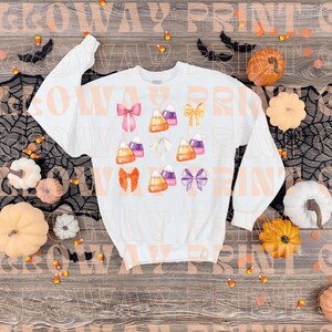 May include: White long-sleeved sweatshirt with a Halloween-themed design featuring candy corn, bows, and ghosts. The design is in shades of orange, purple, and white.