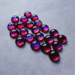 May include: A collection of 20 round glass cabochons with a red and blue iridescent finish. The cabochons are arranged in a triangular shape on a gray surface.