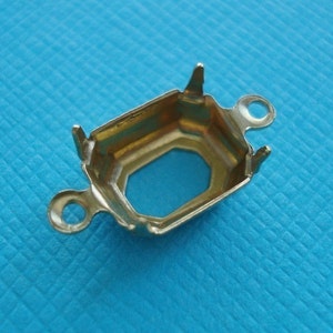 May include: A gold-colored metal jewelry setting with an octagonal center opening. The setting has four prongs and two loops for attaching to other components. The setting is designed to hold a gemstone or other decorative element.