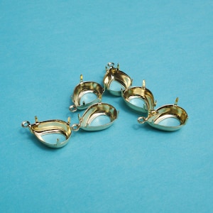 May include: Six gold-tone metal settings for cabochons, each with a teardrop shape. The settings are designed to hold a flat-backed stone or cabochon.