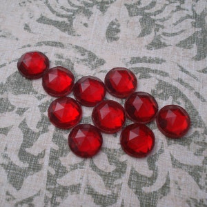 May include: A group of ten red, faceted, glass gems arranged in a staggered pattern on a white background with a floral design.