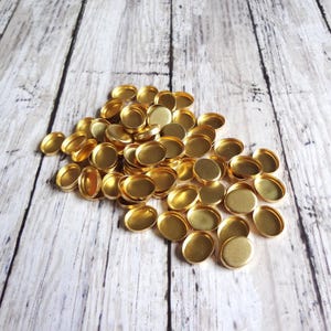 May include: A pile of small, oval-shaped, gold-colored metal bezels. The bezels are empty and have a shallow dish shape. They are scattered on a light-colored, weathered wooden surface.