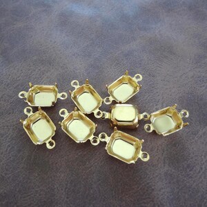 May include: A collection of gold-colored metal jewelry findings. Each finding has a rectangular or square bezel with small prongs and loops on the sides. The findings are scattered on a brown surface.