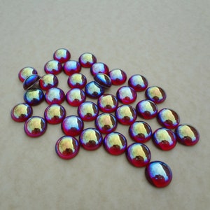 May include: A collection of iridescent glass cabochons in shades of red, blue, and purple. The cabochons are arranged in a loose pile on a beige surface.