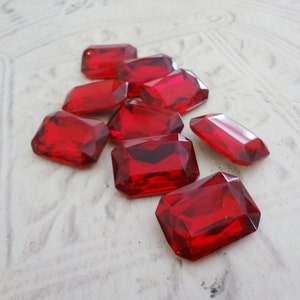 May include: A group of ten red, faceted, rectangular glass gems. The gems are arranged in a loose pile on a white surface.