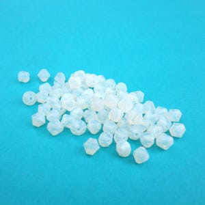 May include: A collection of translucent, white beads with various geometric shapes, including faceted and round designs. The beads are scattered on a bright turquoise background, creating a visually appealing contrast. Ideal for jewelry making and crafting.