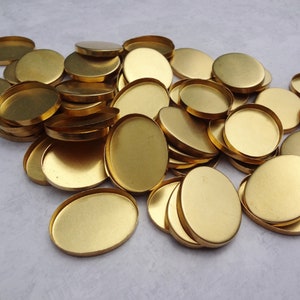 May include: A collection of oval and round brass bezels, ideal for jewelry making or crafting. The bezels are a bright gold color and have a shallow dish shape. They are scattered across a light-colored surface.