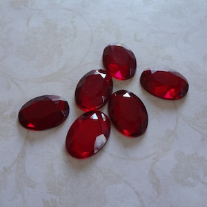 May include: Six oval-shaped, red, faceted glass gems. The gems are arranged in a cluster on a white surface.