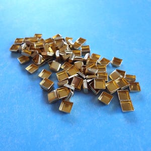 May include: A collection of small, square, gold-toned metal bezels with raised edges. These metallic components feature a hollow center and a smooth, reflective finish, arranged in a scattered pile to display their uniform shape and metallic luster.