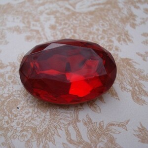 May include: A large, oval-shaped, red glass gem with a faceted design.