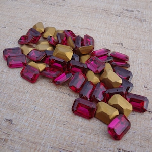 May include: A pile of pink and gold emerald-shaped glass gems. The gems are scattered on a light brown textured surface.