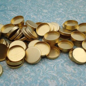 May include: A collection of small, round, brass-colored metal bezels. The bezels have a shallow dish shape with a flat base and a raised rim. They are scattered on a light blue patterned surface, with some stacked and others lying flat.
