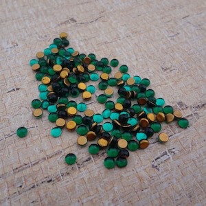 May include: A collection of small, round, emerald green and black glass beads with gold-colored metal settings. The beads are scattered on a light brown, textured surface, creating a visually appealing display of color and texture. These beads are suitable for jewelry making and crafts.