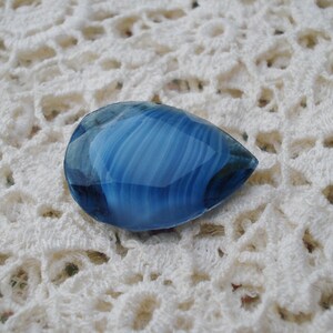 May include: A teardrop-shaped, blue gemstone with white and dark blue striations. The polished stone is set against a white, crocheted background. The gemstone's surface reflects light, highlighting its translucent quality. This could be used for jewelry making.