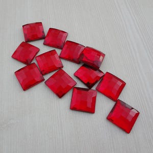 May include: A group of red square-shaped rhinestones, scattered on a light wood surface.