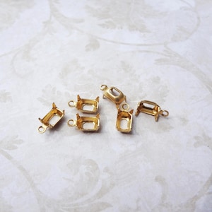 May include: A collection of small, gold-coloured rectangular jewellery settings with prongs and loops. The settings are empty, designed to hold gemstones or other decorative elements. The pieces are scattered on a patterned, off-white surface.