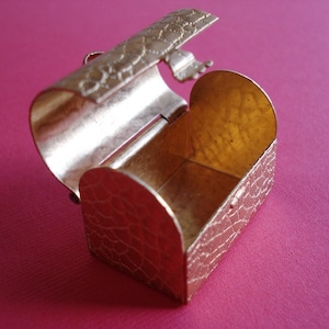 May include: A small, gold-colored metal treasure chest with a hinged lid, open to reveal an empty interior. The chest has a textured, scaly design.
