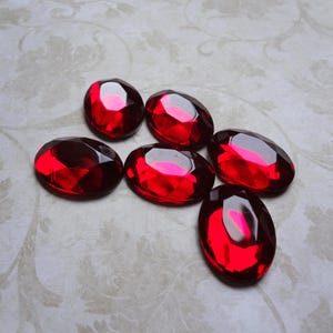 May include: Six oval-shaped, faceted red glass cabochons arranged in a cluster. Each piece features a polished surface with multiple geometric facets that reflect light, creating deep crimson highlights and dark shadows across the smooth, rounded edges.