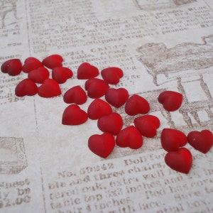 May include: A collection of 20 small, red glass heart-shaped beads scattered on a piece of paper with printed text.