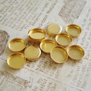 Puede incluir: Una colección de pequeños biseles metálicos dorados y redondos. Los biseles tienen un diseño cóncavo y poco profundo, perfecto para engarzar objetos pequeños. Están colocados sobre un fondo de papel vintage.