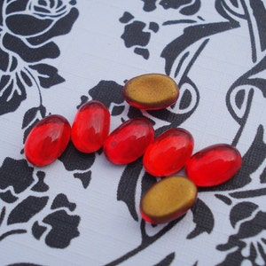 May include: Seven oval-shaped glass beads, six are red and one is gold. The beads are arranged in a curved line on a white surface with a black floral pattern.
