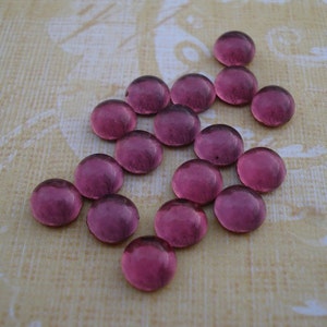 May include: A collection of 16 round, translucent, purple glass beads. The beads are arranged in a loose, asymmetrical pattern on a beige fabric background.