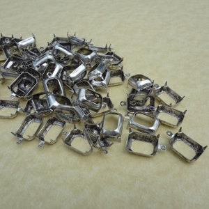 May include: A collection of silver-colored metal jewelry findings, specifically bezel settings. The settings are rectangular with prongs and small loops for attaching to other components. The findings are scattered on a light beige surface.