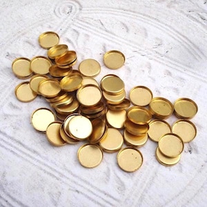 May include: A collection of small, round, brass-colored metal bezels. The bezels are scattered across a white surface with a subtle wood grain pattern. Each bezel has a shallow, circular indentation.