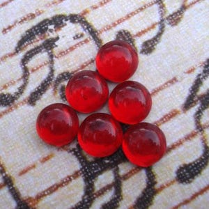 May include: Six small, round, translucent red glass beads are arranged on a patterned, off-white surface. The beads have a glossy finish and reflect light, creating highlights. The background features a faded musical score.