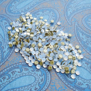 May include: A pile of small, round, white and gold-rimmed embellishments. The embellishments are scattered on a blue and brown paisley patterned fabric.