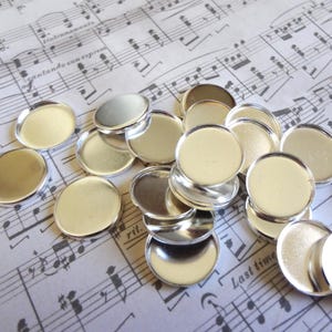 May include: A collection of silver-colored, round bezel trays, perfect for jewelry making or crafting. The trays are scattered on a sheet of paper with musical notes printed on it. The trays have a flat surface and a raised edge.